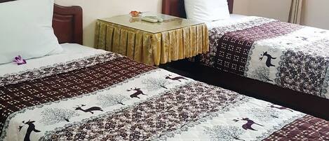 A hotel room with two beds, each with a patterned bedspread, a small table with a lamp, and a window with curtains.