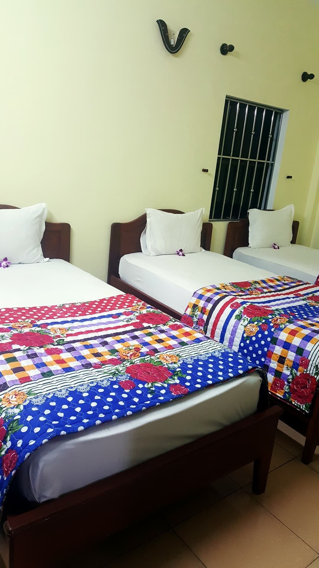 A hotel room with two single beds, each with a colorful quilt and white pillows. The room has a simple layout with a window and a door visible.