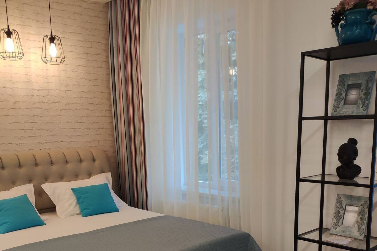 A bedroom with a large bed, a black metal shelf with decorative items, and a window with sheer curtains.