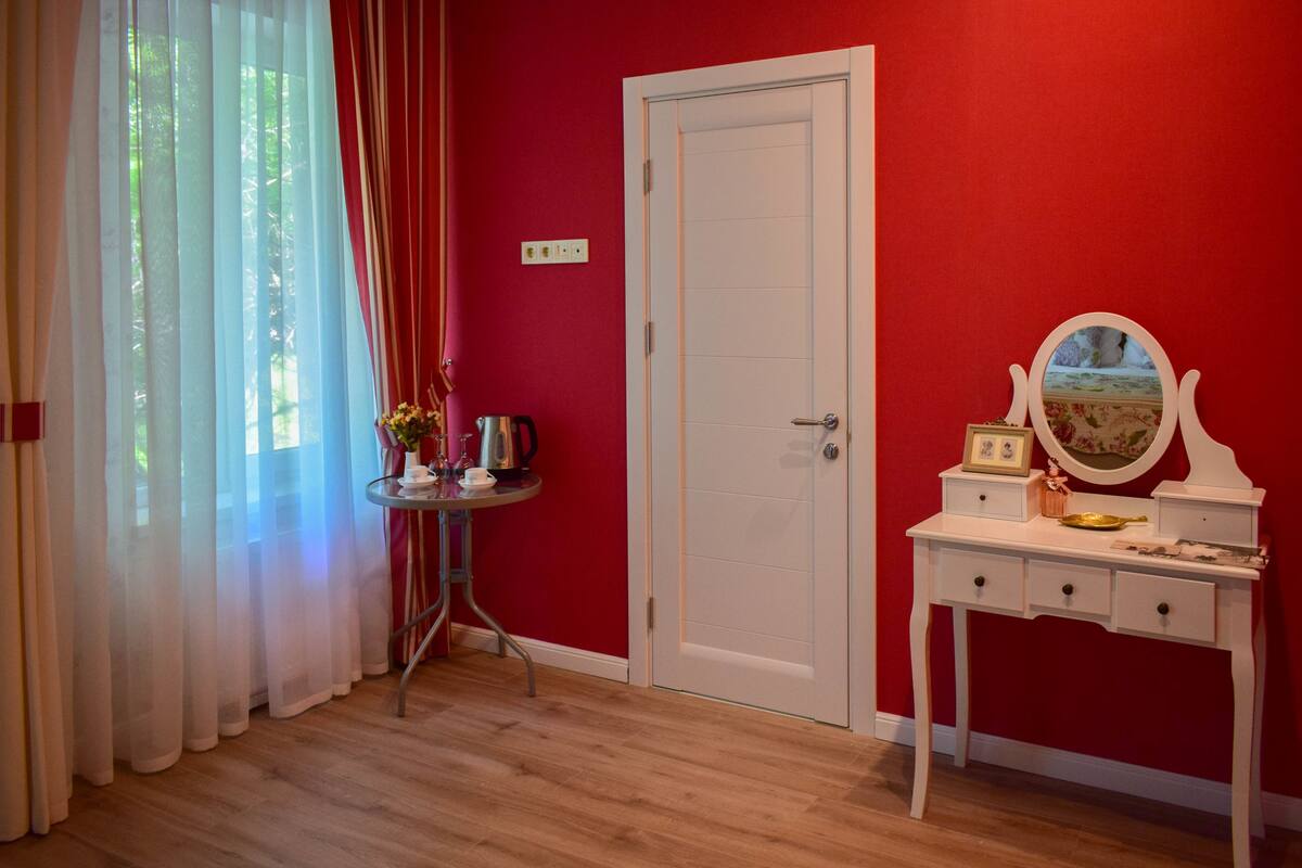 A room with a red wall, a white door, a small table with a kettle, and a vanity with a mirror.