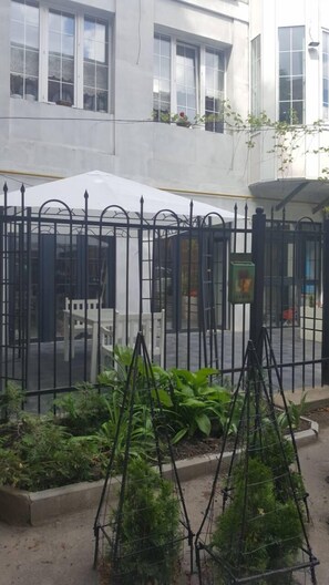 Un patio amurallado con un edificio blanco al fondo, una cerca de metal negro y algo de vegetación.
