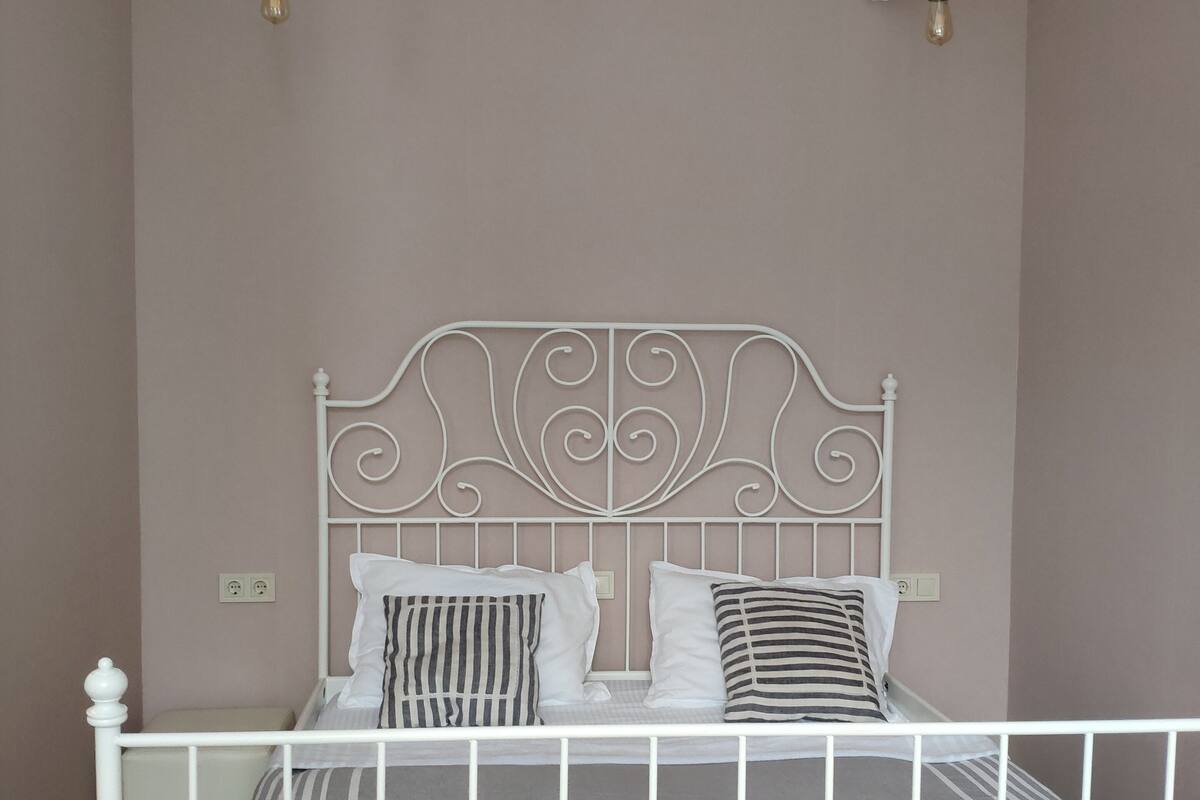 A white metal bed frame with a decorative headboard, two pillows, and a striped bedspread in a room with light pink walls and wooden flooring.