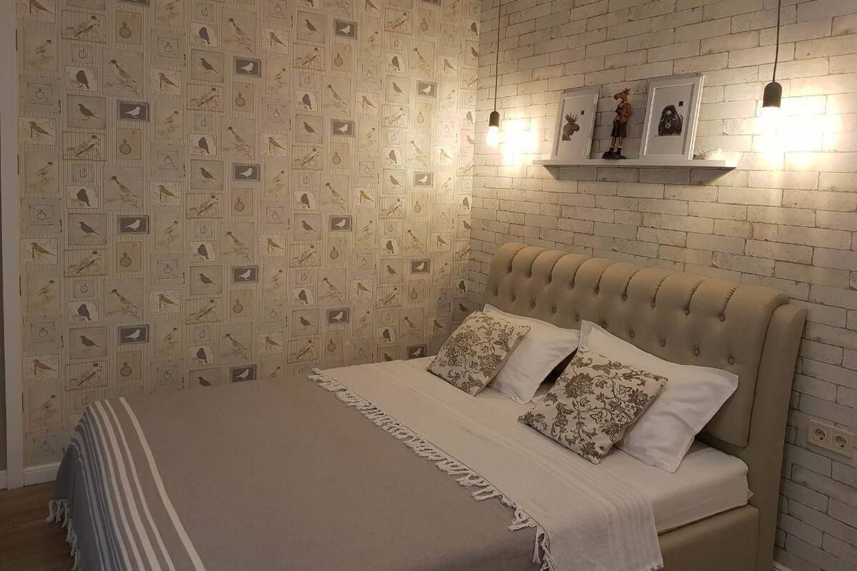 A bedroom with a patterned wallpaper, a tufted headboard, and a bedside shelf with framed pictures.