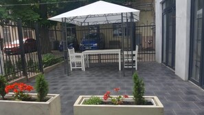 Un patio al aire libre con una mesa y sillas, rodeado por una valla metálica y un paraguas blanco.