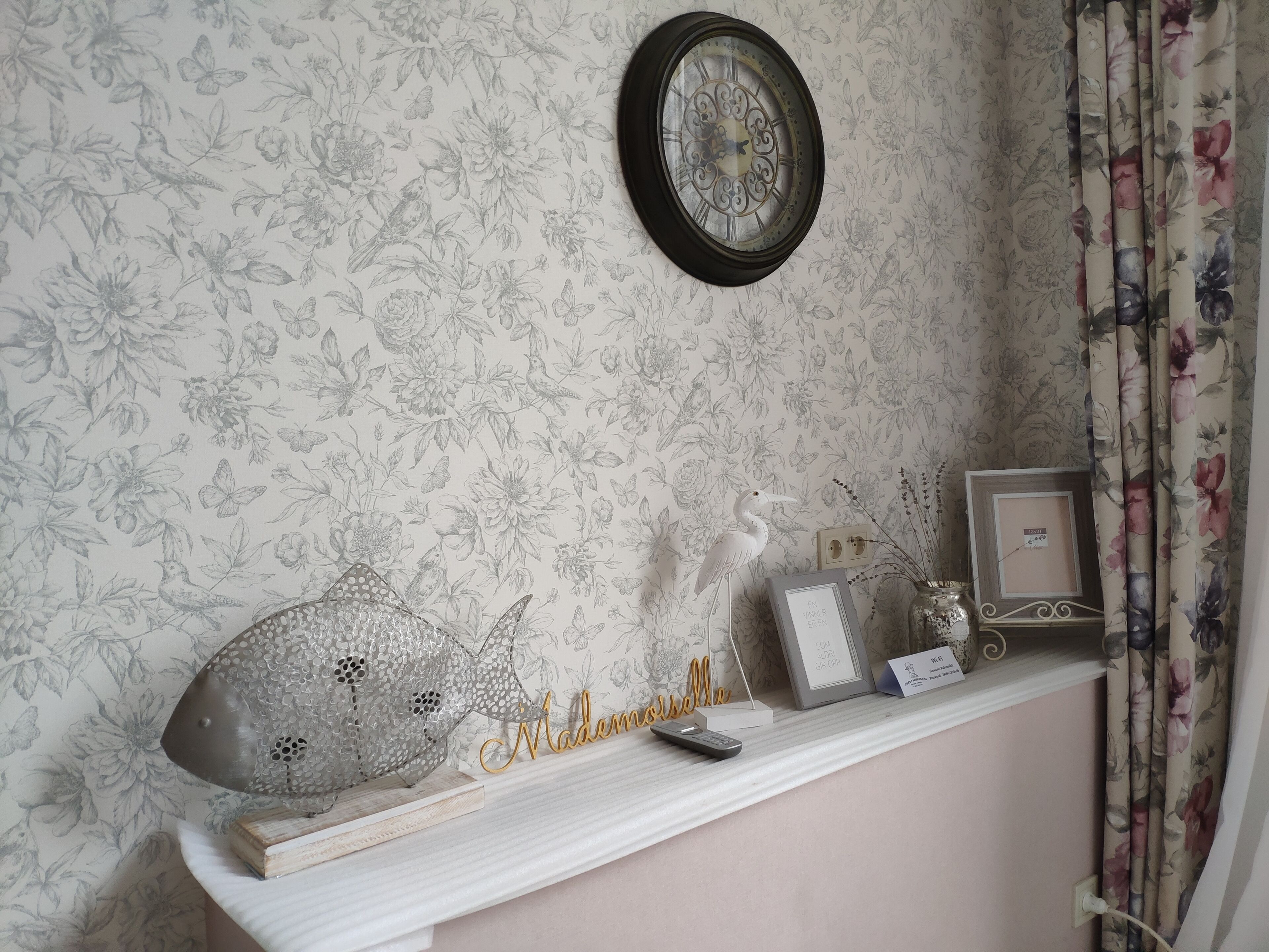 A wall with a floral wallpaper, a clock, a shelf with decorative items, and a framed picture.