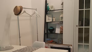 Una habitación con una mesa blanca, dos sillas, una lámpara de pie, una estantería con objetos decorativos y una puerta con paneles de vidrio.