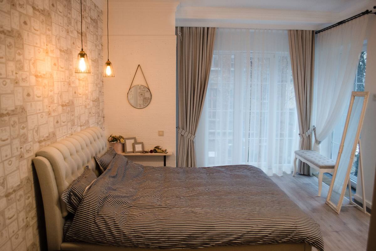 A bedroom with a brick-patterned wall, a bed with striped bedding, two hanging light fixtures, a bedside table with a mirror, and a window with sheer curtains.