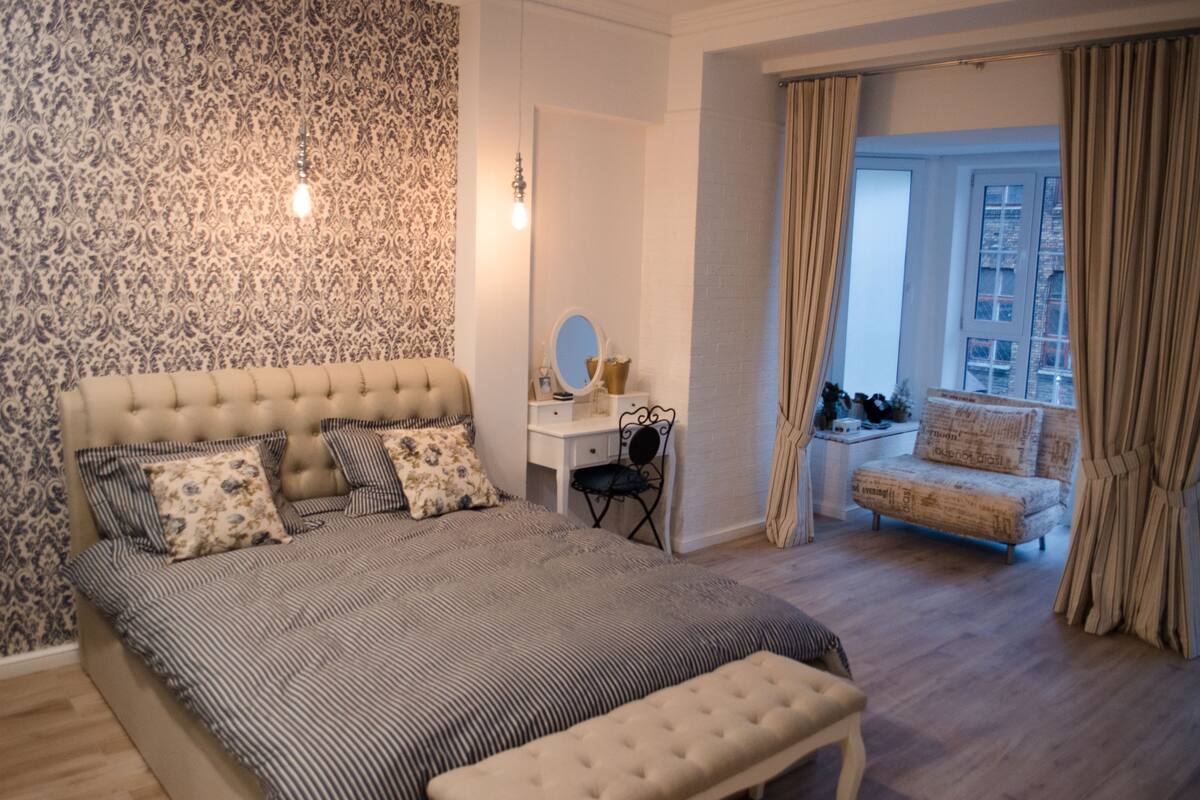 A bedroom with a large bed, a desk, and a chair. There is a window with curtains and a view of buildings outside.