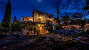 Una casa de piedra con balcón, rodeada de cactus y árboles, bajo un cielo nocturno con luna visible.