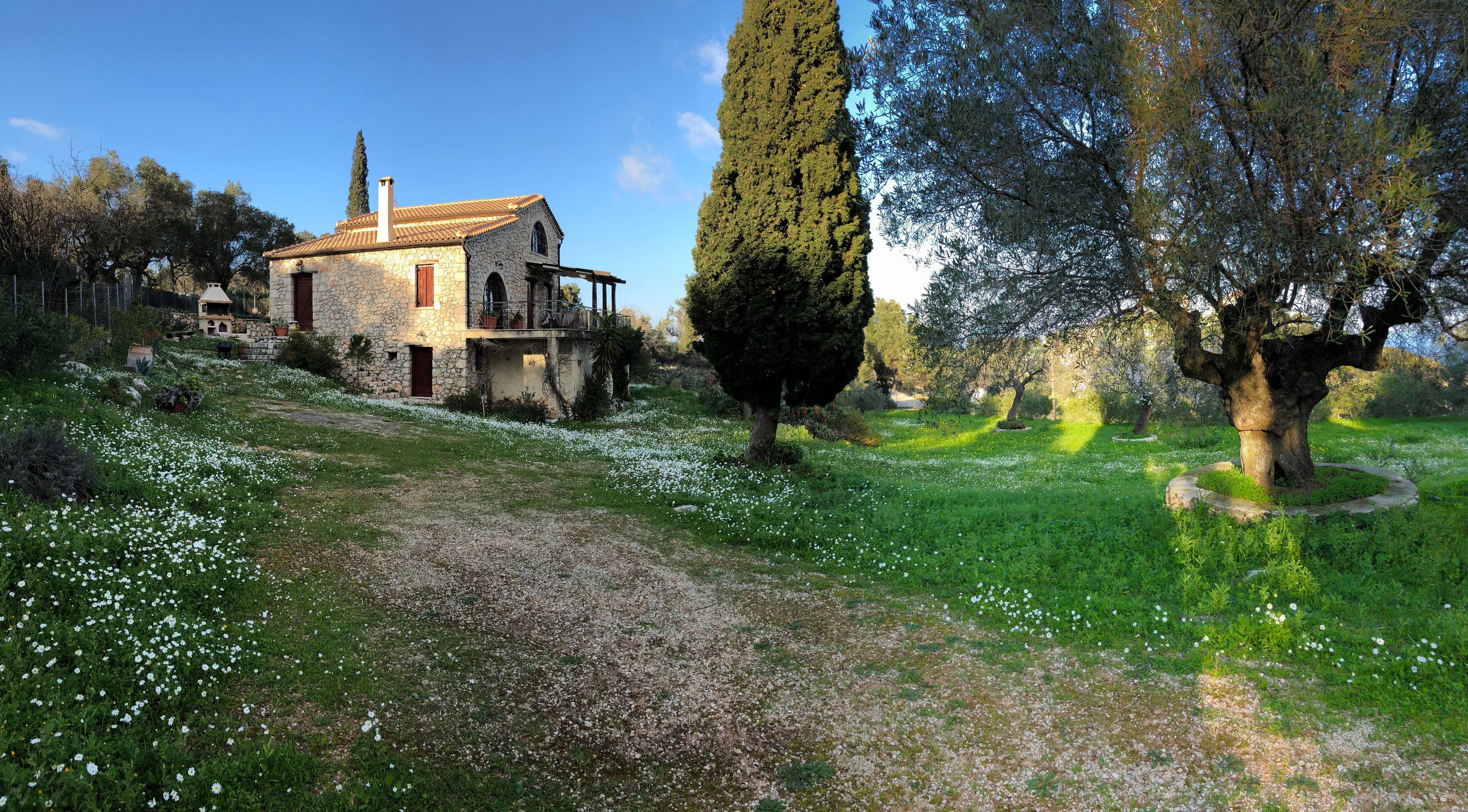 A stone house with a balcony, surrounded by a garden with white flowers and tall trees.