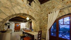 Cocina rústica de piedra con una mesa y sillas de madera, un arco de piedra y vistas a través de una ventana con celosías de madera.