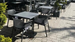 Un café al aire libre con mesas y sillas, una zona de estar cubierta y un edificio con contraventanas verdes al fondo.
