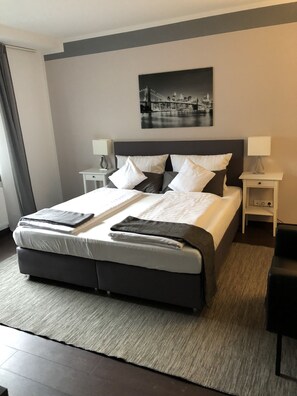 A neatly made bed with a dark headboard, two bedside tables with lamps, a black and white cityscape painting above the bed, and a dark floor. - Hotel Restaurant Zum alten Postwagen (Gevelsberg)