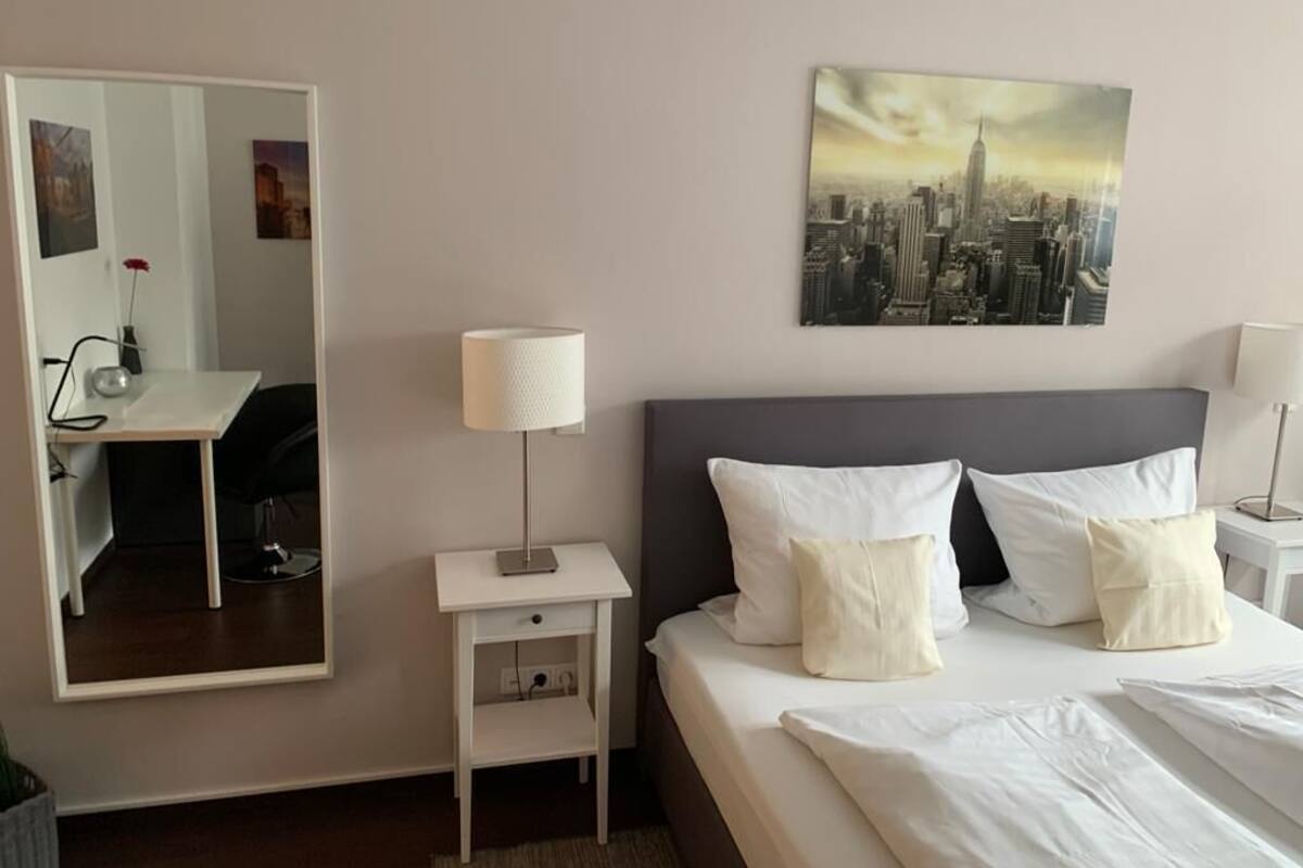 Ett hotellrum med en säng, ett skrivbord, en stol, en lampa och en tavla med en stadsbild på väggen.