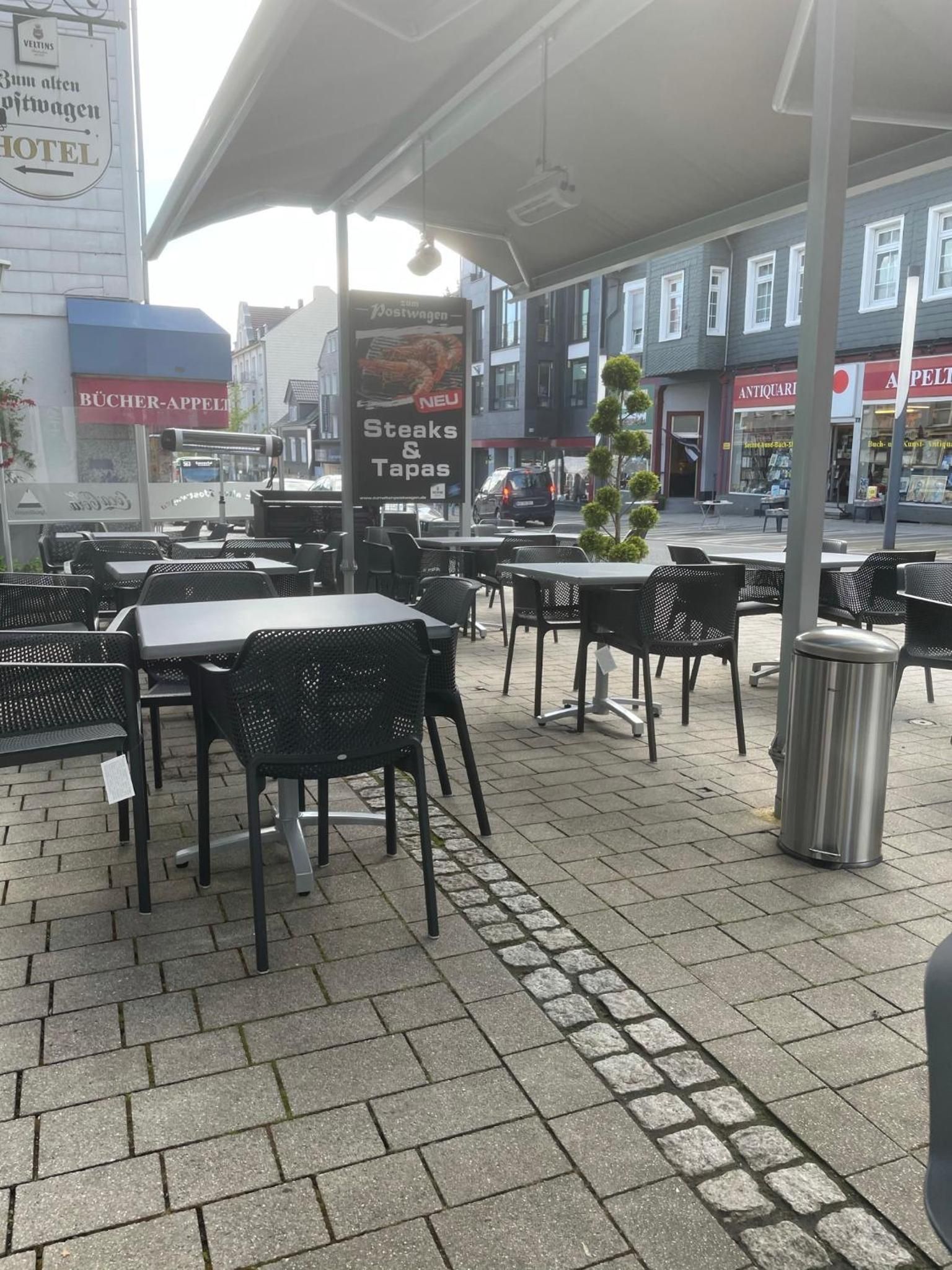 An outdoor seating area with tables and chairs, a trash can, and a sign advertising steaks and tapas.