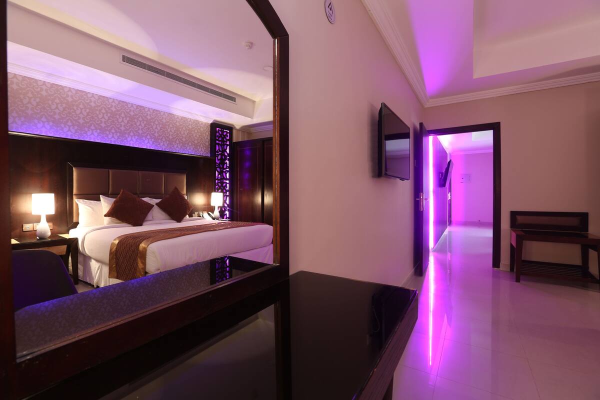 A hotel room with a bed, bedside table, lamp, and a mirror reflecting the room.
