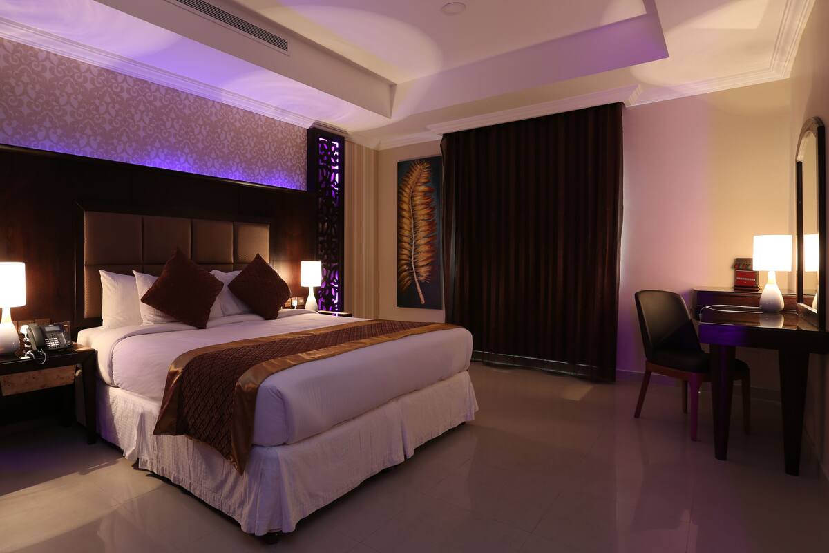 A hotel room with a large bed, a desk with a chair, a nightstand with a lamp, and a decorative wall panel.