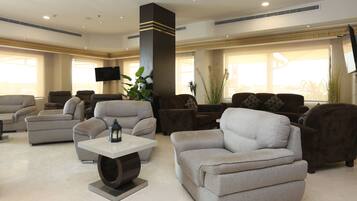 A spacious living room with multiple sofas and armchairs, a flat-screen TV mounted on the wall, and large windows providing natural light.