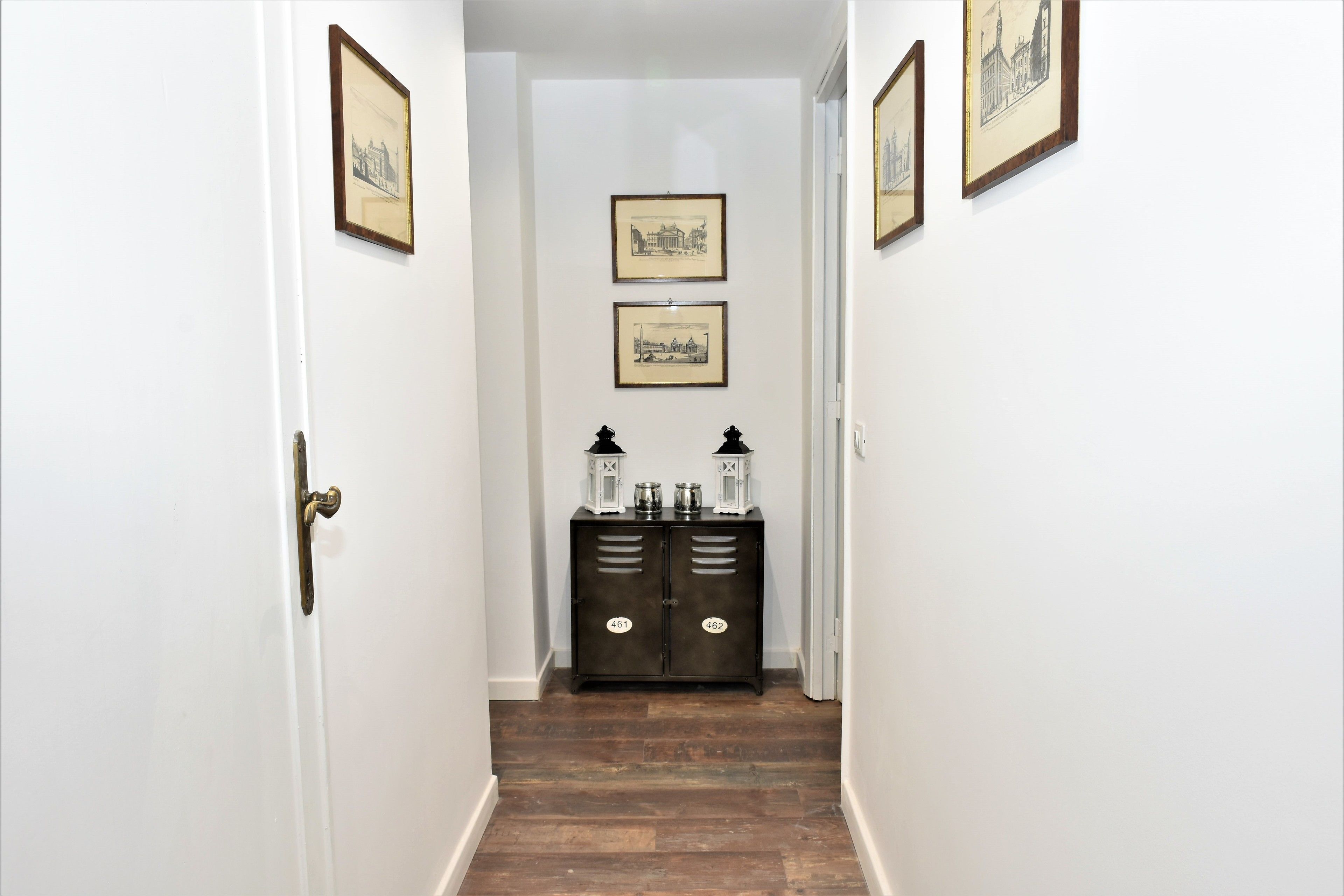 A hallway with wooden flooring, white walls, and two framed pictures on the wall. There is a dark-colored cabinet with two white lanterns on top.