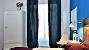 A bedroom with a bed, a nightstand, a lamp, a chair, a wardrobe, and a window with curtains. - Coliseum Suite (Rome)