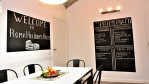 A dining area with a table set for two, a fruit bowl, and a wall sign listing Italian dishes. - Coliseum Suite (Rome)