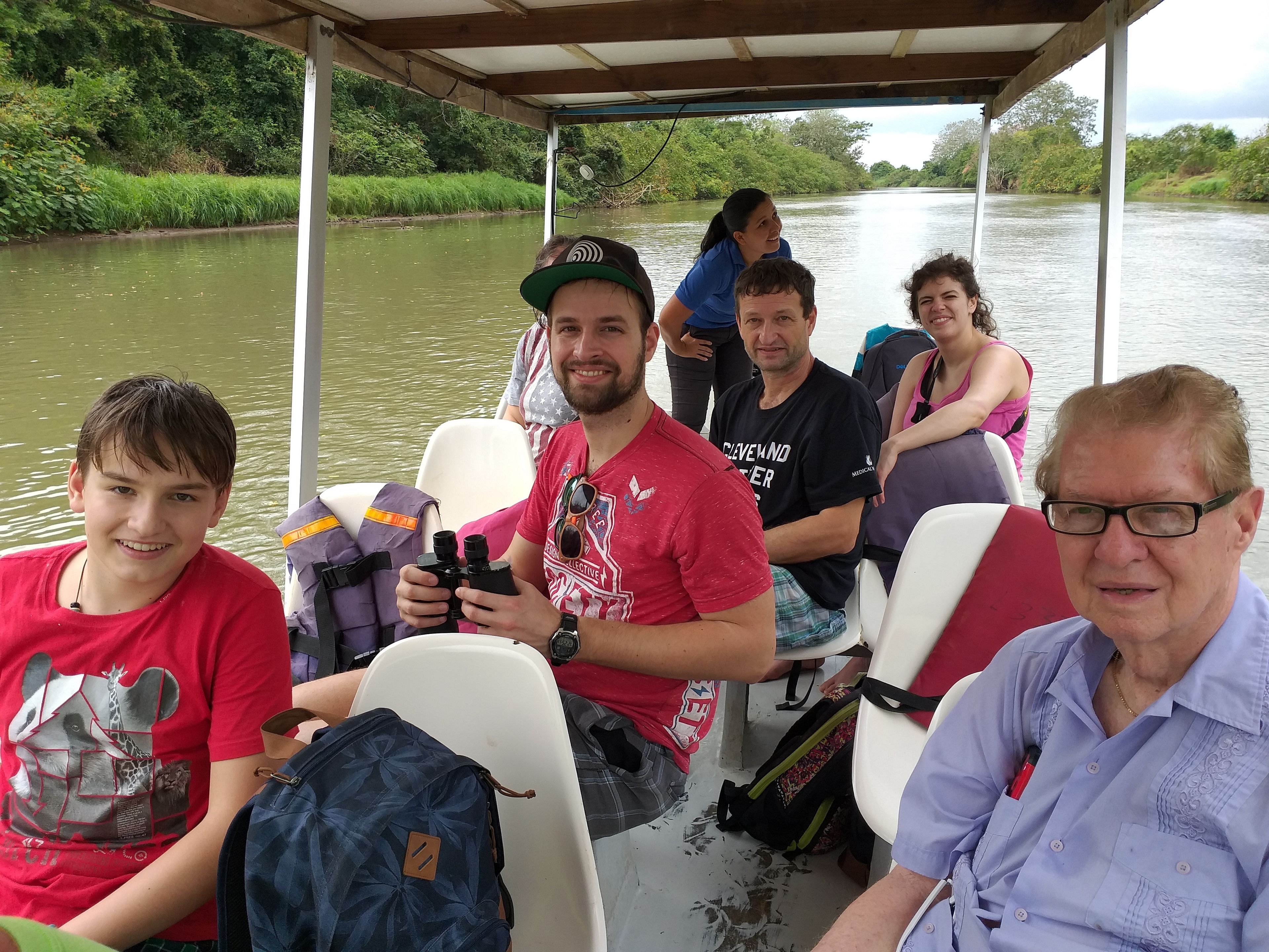 Um grupo de pessoas em um barco, com toldo sobre a cabeça, cercado por água e vegetação.