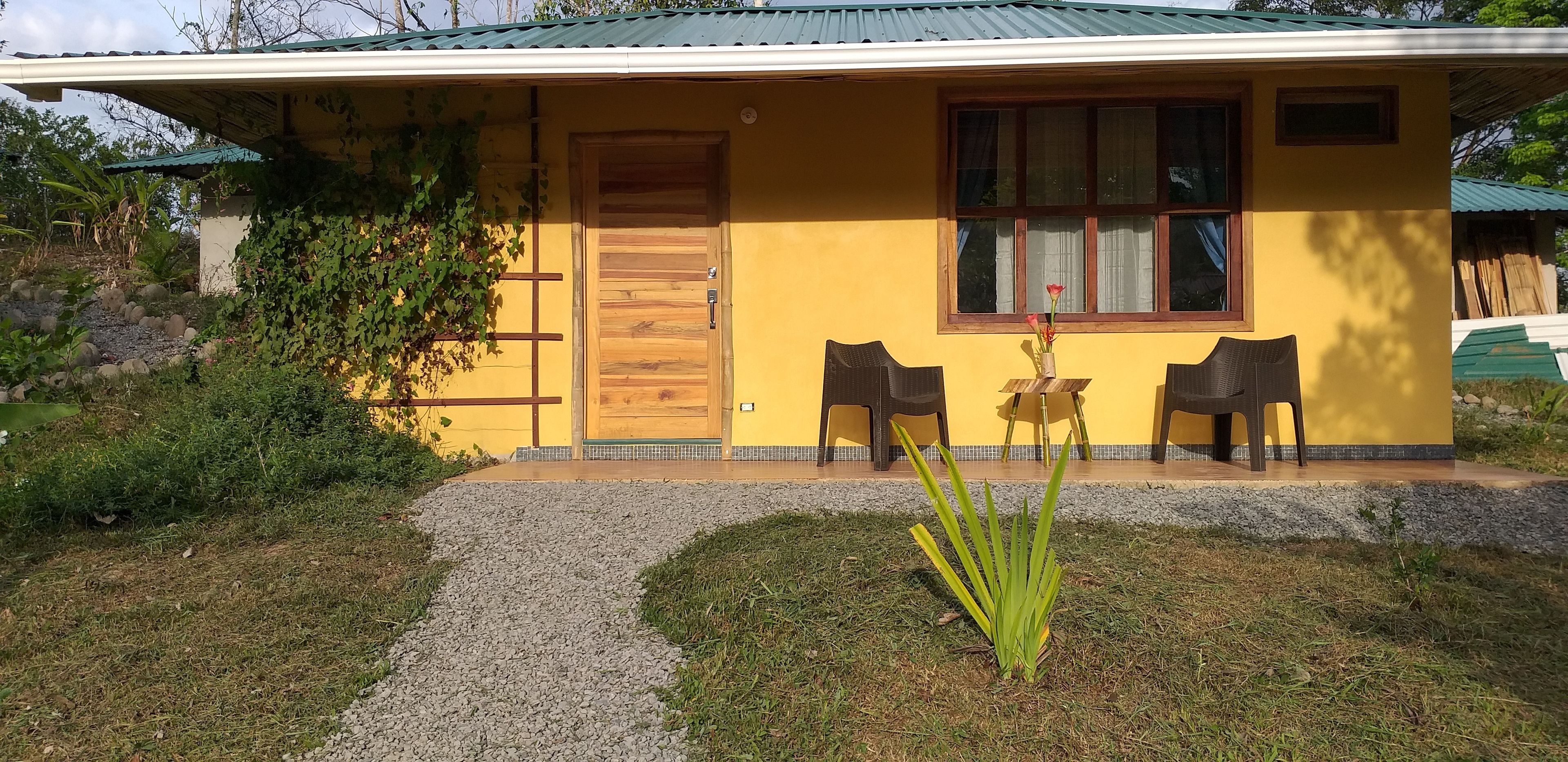 A yellow house with a wooden door, two chairs outside, and a small plant in front.