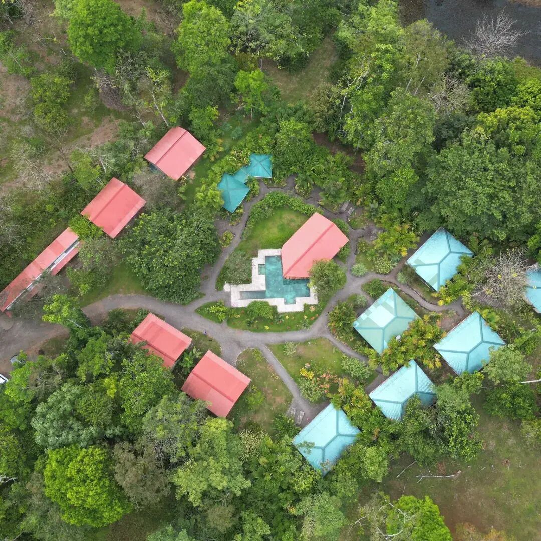 An aerial view of a resort with multiple buildings, a central swimming pool, and surrounding lush greenery.