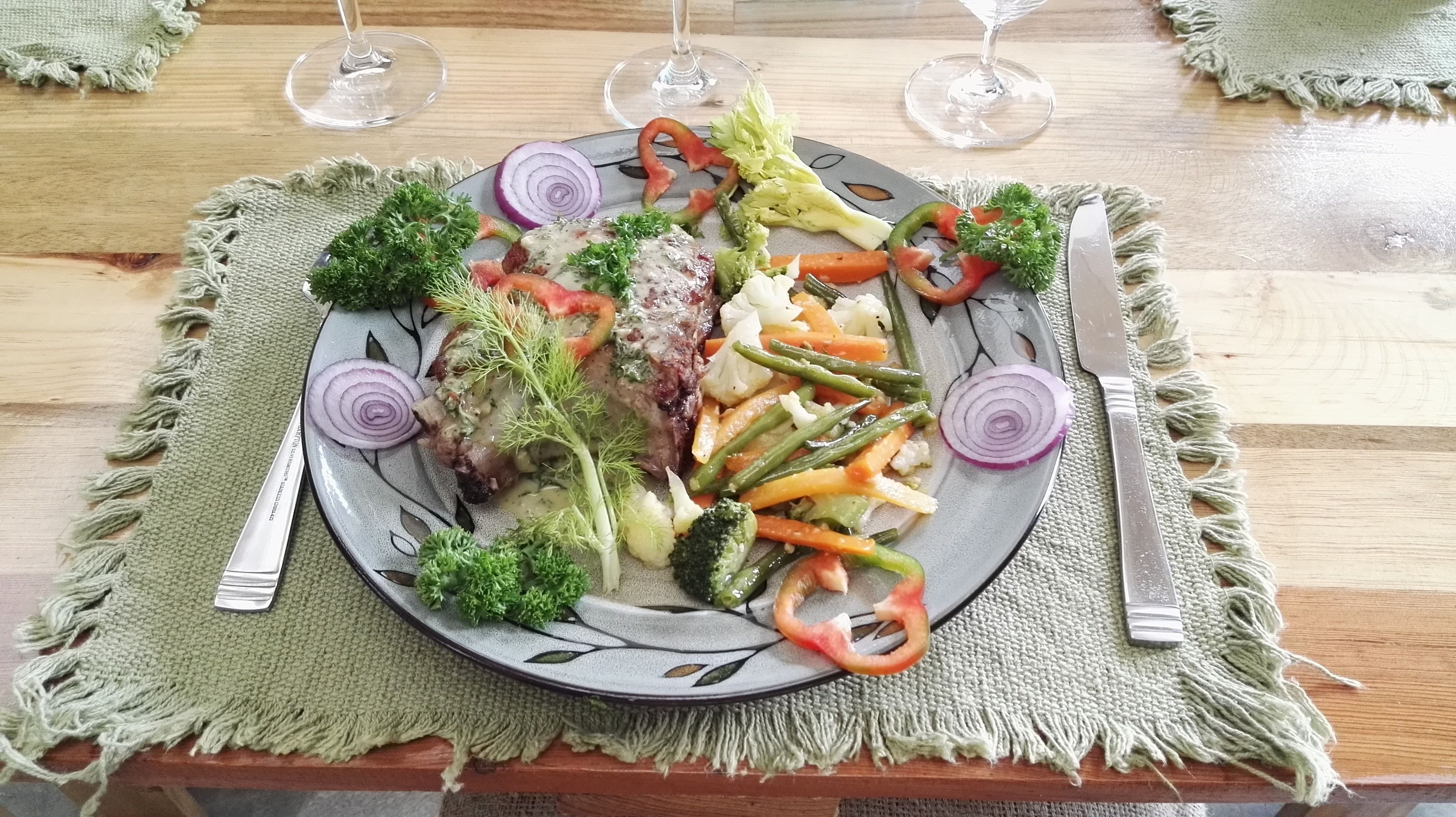 A plate of food with vegetables and a steak, accompanied by a knife and fork, on a table with a woven placemat.