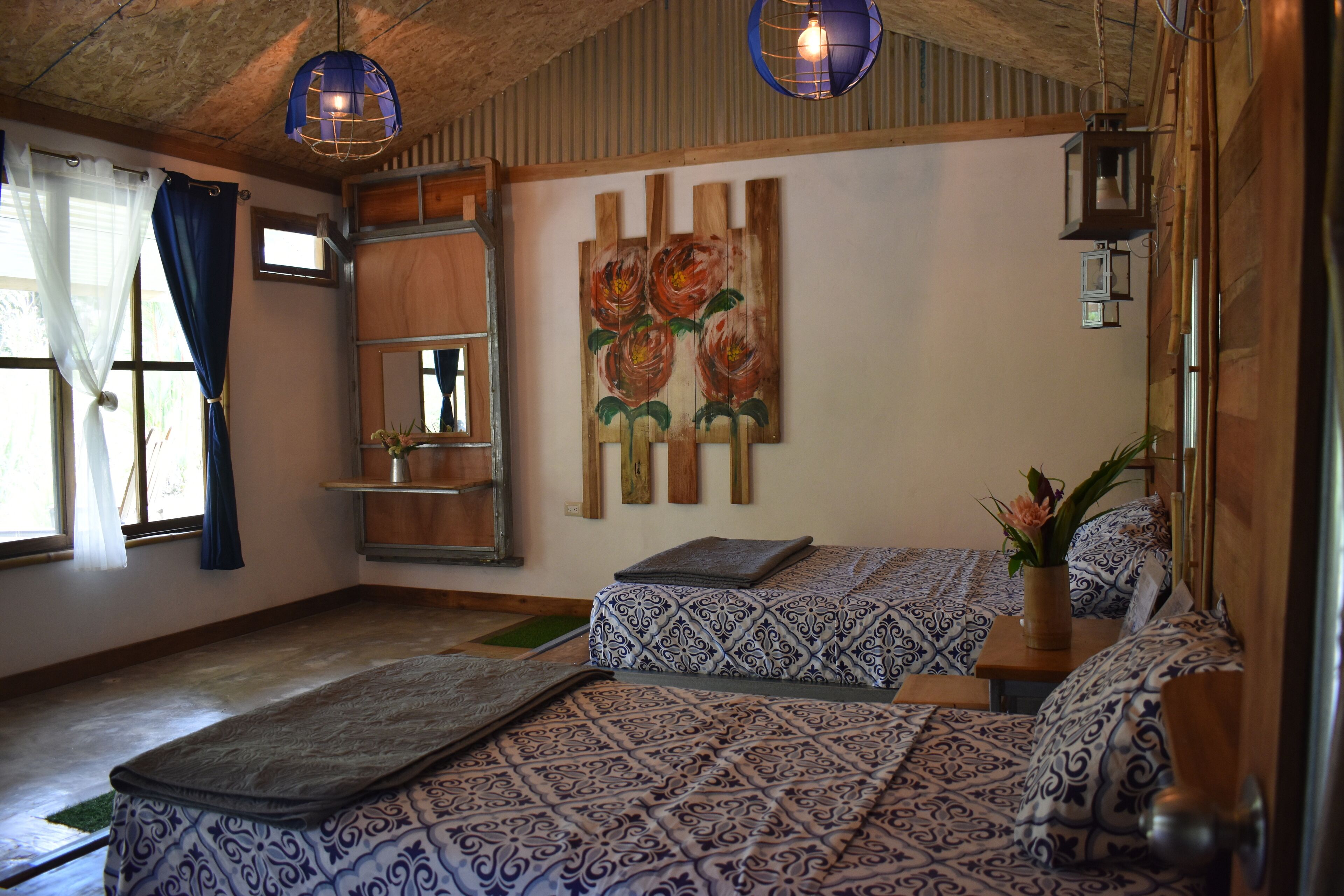 A room with two beds, a wooden ceiling, a painting of flowers, and a small shelf with a vase.