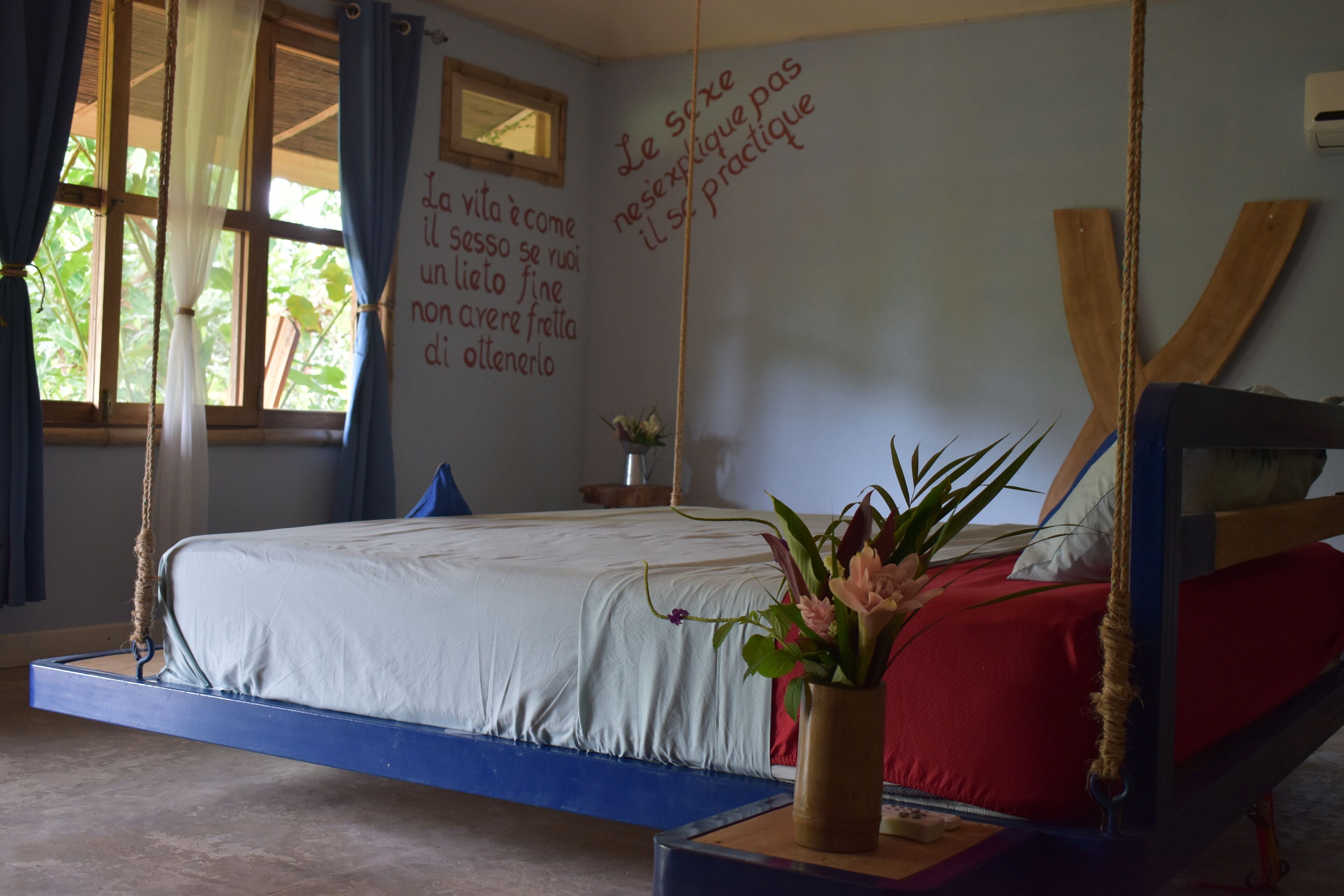 A bedroom with a bed, a wall with a quote, a window, and a hanging plant.