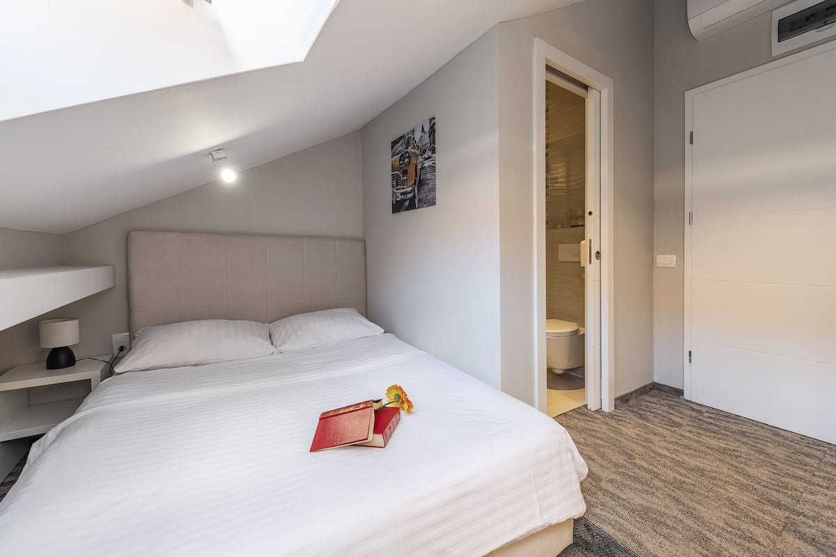 Ein Schlafzimmer mit Bett, Nachttisch, Wandregal, Tür und Deckenleuchte.