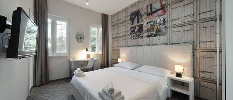 Una habitación de hotel con una cama grande, dos mesitas de noche con lámparas, un escritorio con una silla, un televisor y una pared decorada con carteles de paisajes urbanos.