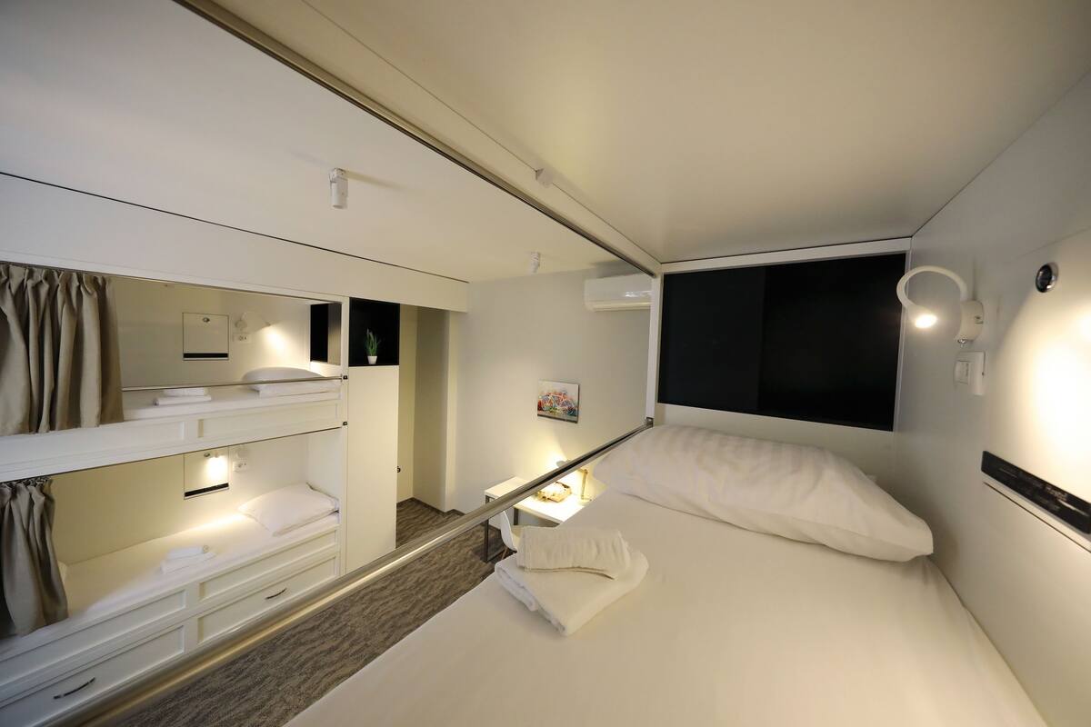 Ein Hotelzimmer mit Etagenbett, einem Einzelbett, einem Nachttisch mit Lampe, einer Wand-Klimaanlage und einem Fenster mit Verdunkelung.