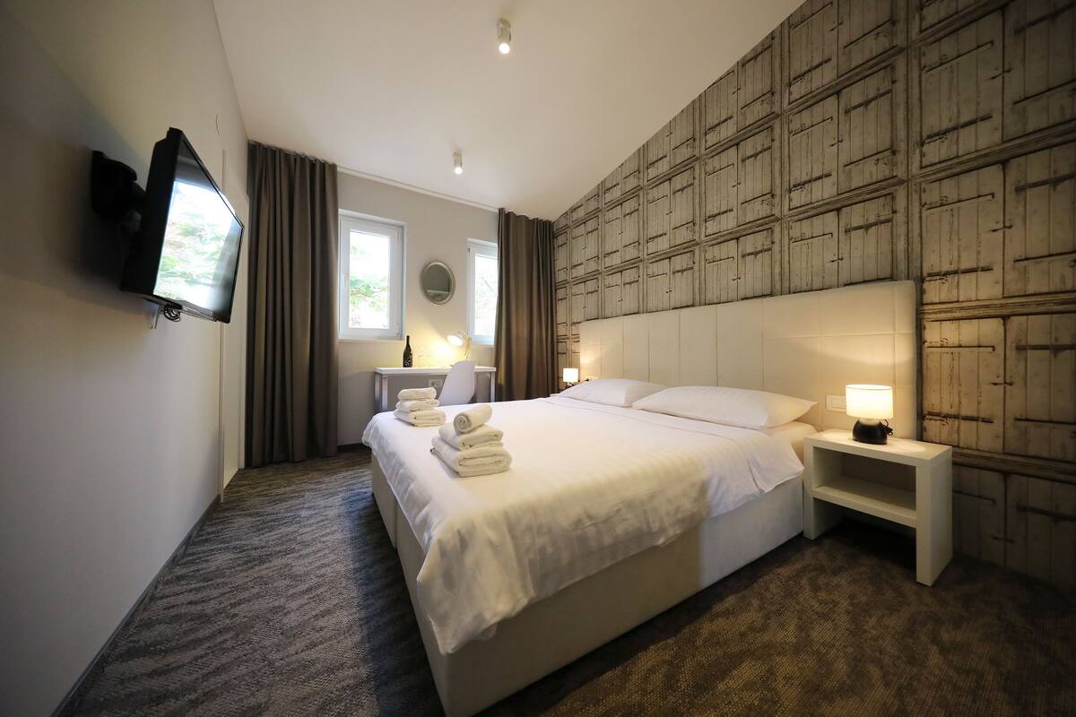 Ein Hotelzimmer mit einem großen Bett, einem flachbildfernseher an der Wand, einem Nachttisch mit einer Lampe und einer Wandverkleidung aus Holz.