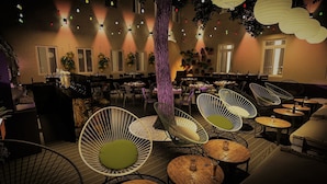 Un restaurante acogedor con mesas redondas y sillas de mimbre, luces colgantes y plantas en macetas.