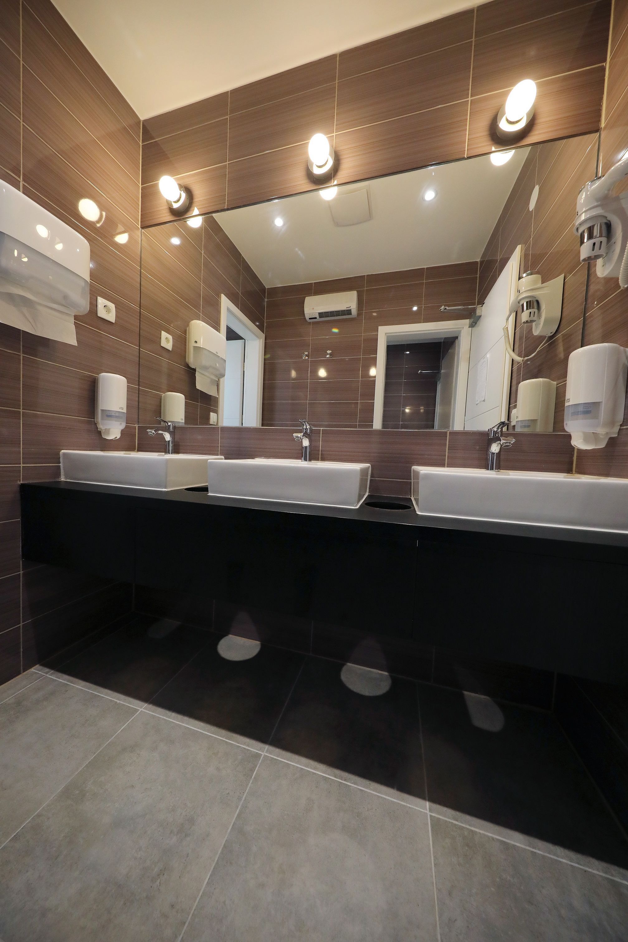 Une salle de bain moderne avec deux lavabos, un grand miroir et des sèche-mains fixés au mur.