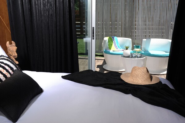 A hotel room with a bed, two white wicker chairs, a small table with a drink, and a straw hat on the bed.
