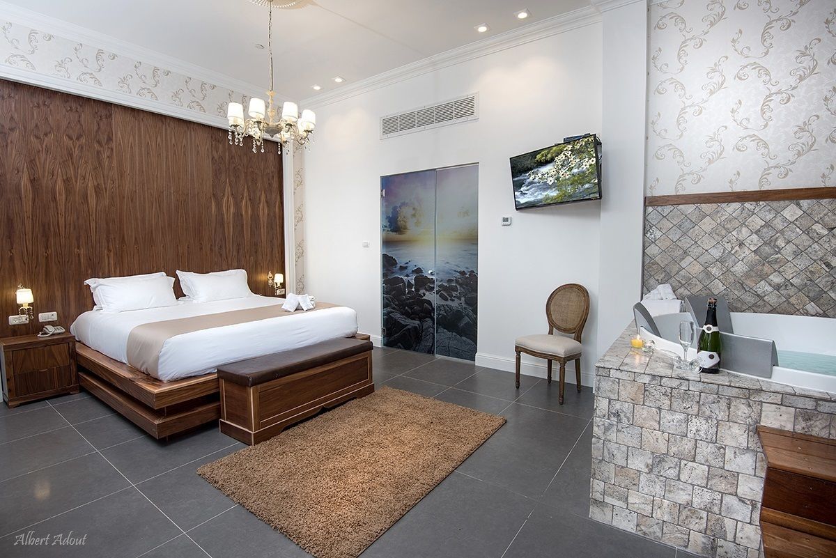 A modern bedroom with a large bed, a wooden headboard, a TV mounted on the wall, a small seating area, and a stone-tiled bathroom with a bathtub.