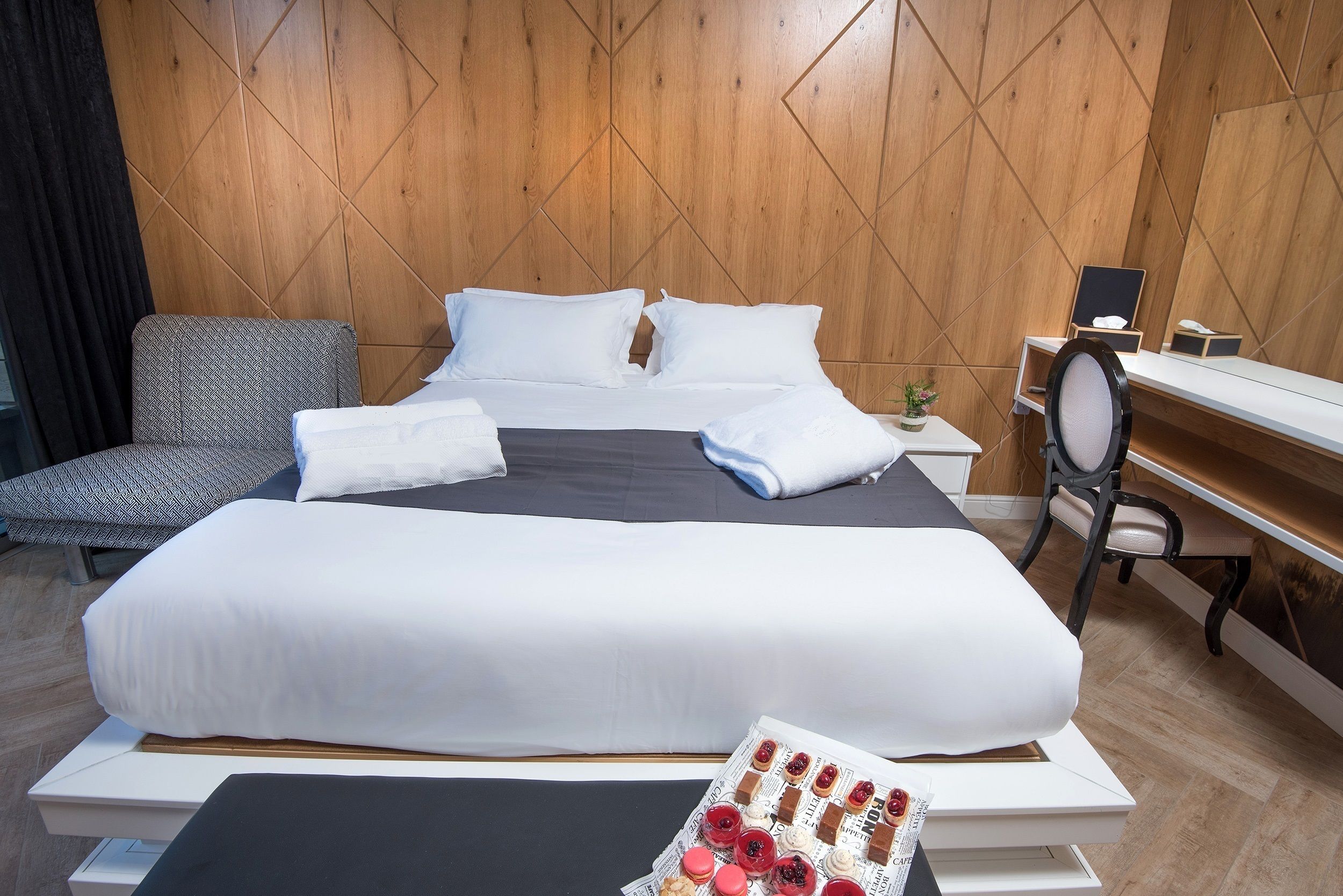A hotel room with a large bed, a desk, a chair, and a small table with a tray of pastries.