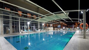 Ein Swimmingpool mit hellem, blauen Fliesenboden, umgeben von Liegestühlen und Sonnenschirmen, unter einem Vordach mit modernem architektonischen Design.