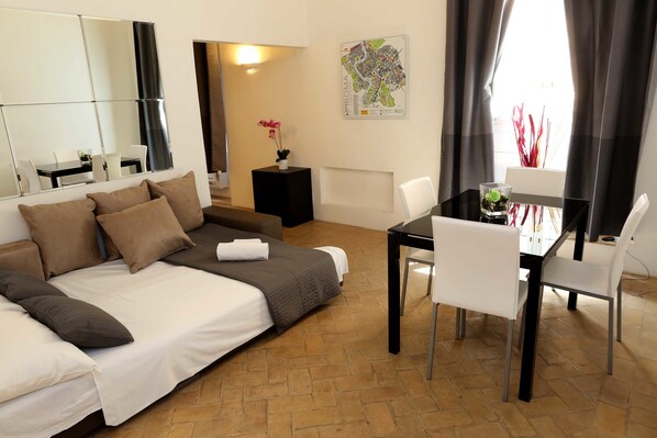 Ein Hotelzimmer mit Bett, einem Esstisch mit Stühlen, einem kleinen Beistelltisch mit Vase, einer Karte an der Wand und einem großen Spiegel.