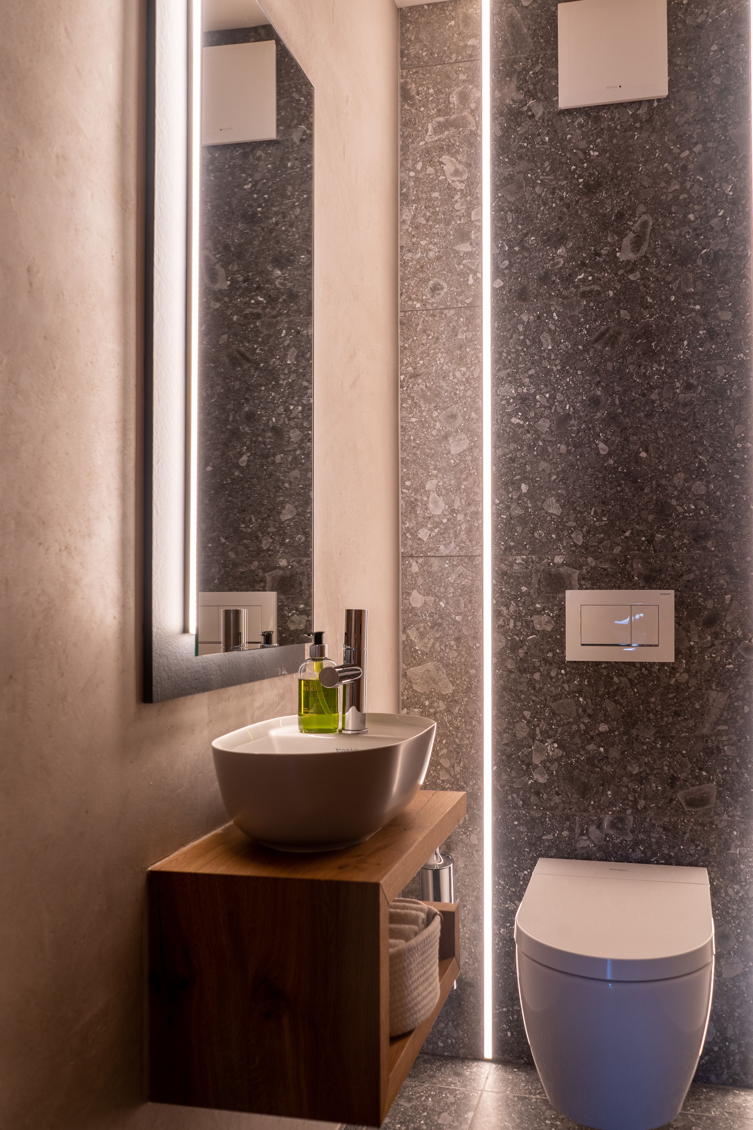 A modern bathroom with a marble sink, a wooden vanity, a toilet, and a wall-mounted light fixture.