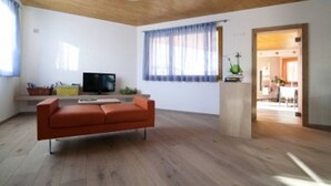 A modern living room with a wooden ceiling, a sofa, a television, and a window with curtains. - Agritur I Colori (Sanzeno)