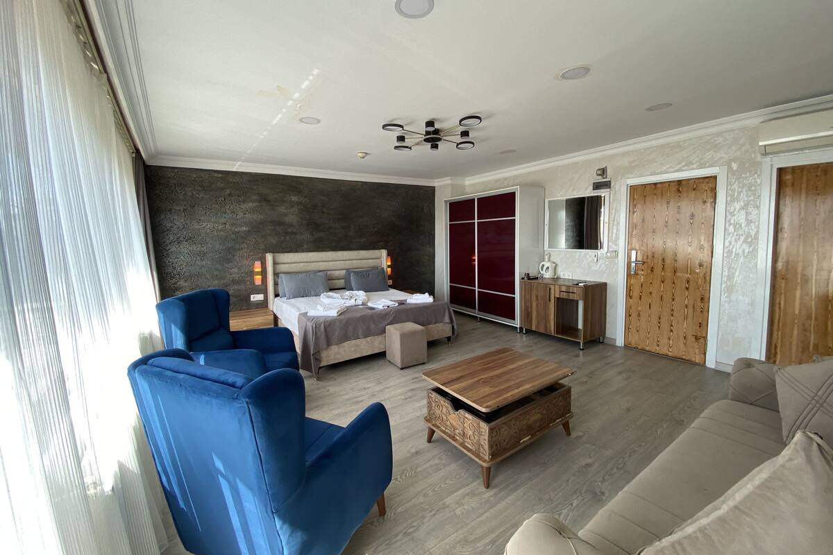 Quarto de hotel moderno com cama, escrivaninha, poltrona azul, mesa de centro de madeira e sofá bege.