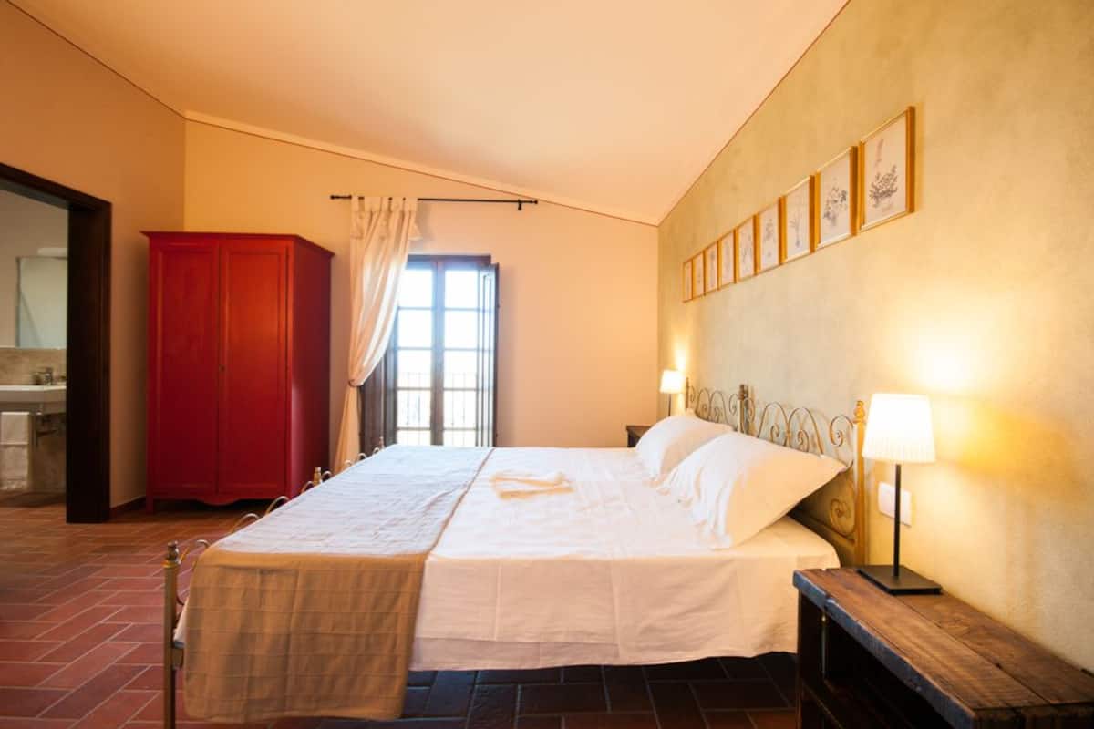A bedroom with a bed, bedside lamps, a red wardrobe, a window with curtains, and framed pictures on the wall.