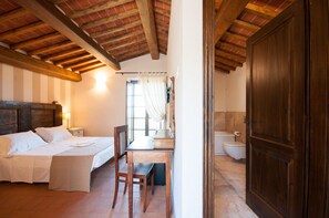 A bedroom with a bed, bedside table, and a wooden chair. There is a window with curtains, a wooden door, and a bathroom with a toilet and sink. - Borgo tra gli Olivi (Castiglion Fiorentino)