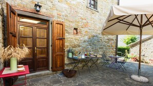 A stone building with a wooden door, a table with chairs, and a table umbrella. - Borgo tra gli Olivi (Castiglion Fiorentino)
