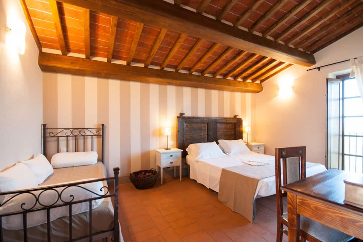 A room with two beds, a wooden ceiling, a window with curtains, and a table with a book.