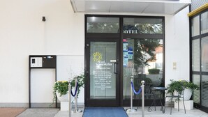 Een hotelentree met een blauw bord, een receptiebalie, een blauwe mat, potplanten en een glazen deur.